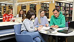 Studierende (divers) sitzen an Tischen in der Bibliothek und lernen gemeinsam. Im Hintergrund sind Bücherregale zu sehen.